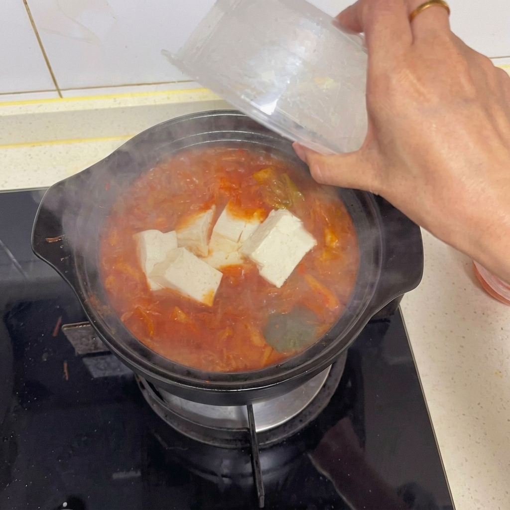 끓고 있는 빨간 김치찌개 냄비에 투명한 플라스틱 용기에서 하얗고 큼직하게 썬 두부를 조심스럽게 넣는 손.