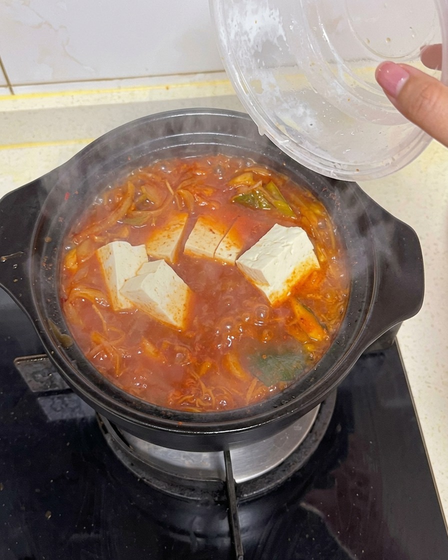 채소와 다시마가 들어간 진한 빨간색 김치찌개가 끓고 있는 냄비에 하얗고 네모난 두부 조각들을 넣는 모습.