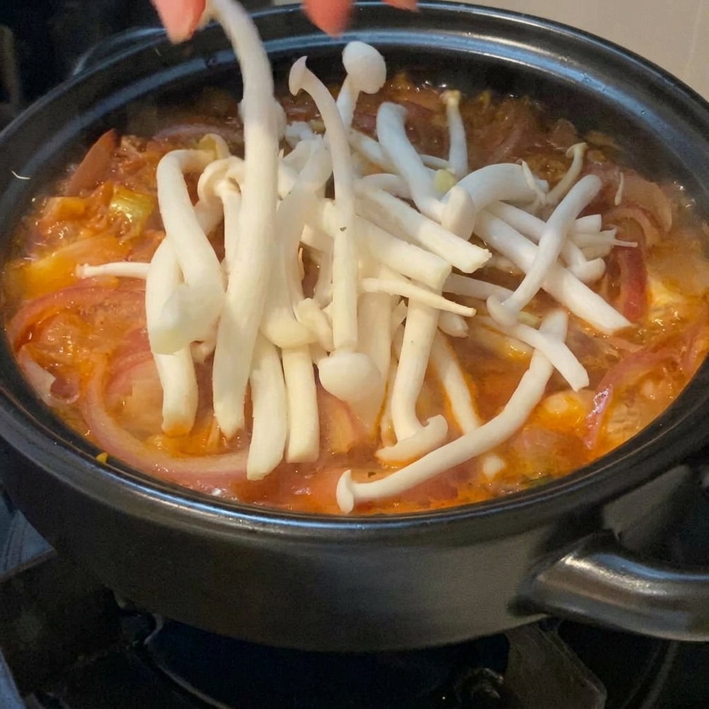 검은색 뚝배기 안에서 끓고 있는 붉은 김치찌개 위에 신선한 흰색 백만송이버섯을 손으로 조심스럽게 올리는 모습.