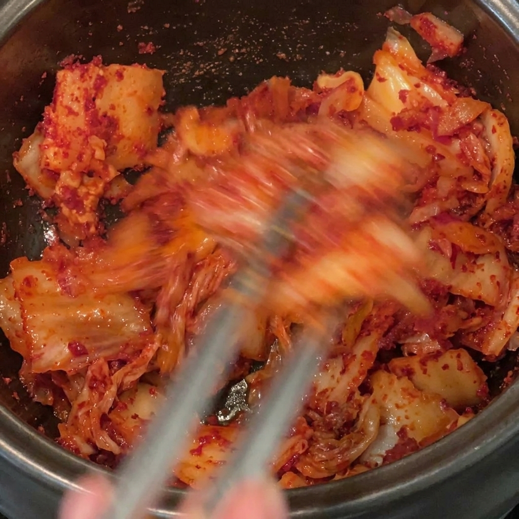 검은 냄비 안에서 김치, 삼겹살, 고춧가루를 골고루 섞으며 볶는 모습.