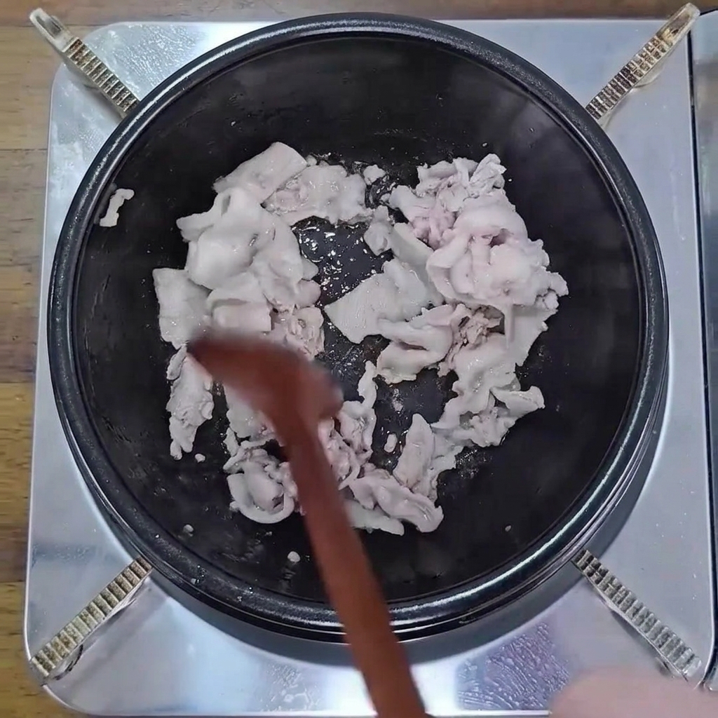 검은 냄비 안에서 나무 숟가락으로 얇게 썬 삼겹살을 노릇하게 볶는 모습.