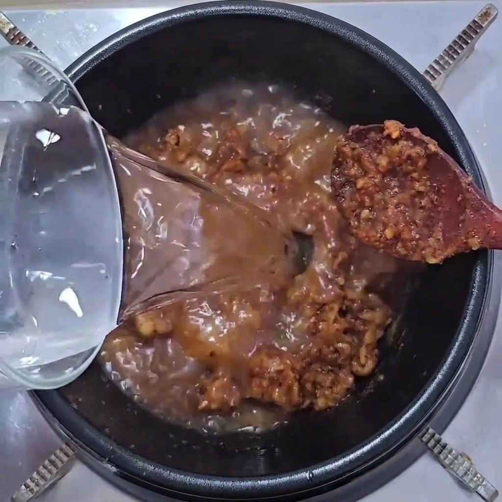 돼지고기와 된장 양념이 지글지글 끓고 있는 냄비에 유리그릇으로 물을 붓는 모습.