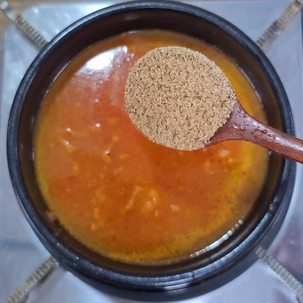 붉은빛이 도는 찌개 국물 위로 소고기 다시다 가루가 담긴 나무 숟가락을 들고 있는 모습.