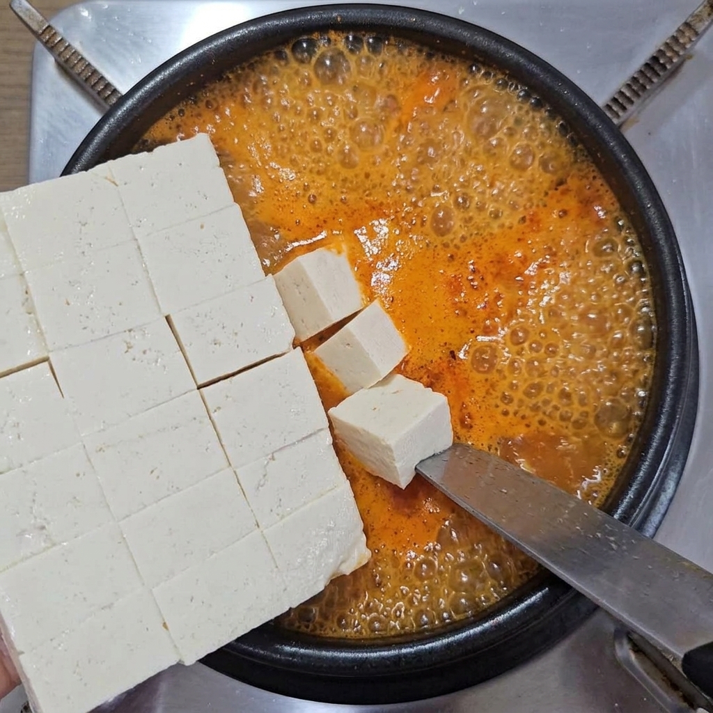 깍둑썰기한 흰 두부를 큰 칼에서 미끄러뜨리듯 끓는 찌개 냄비에 넣는 모습.