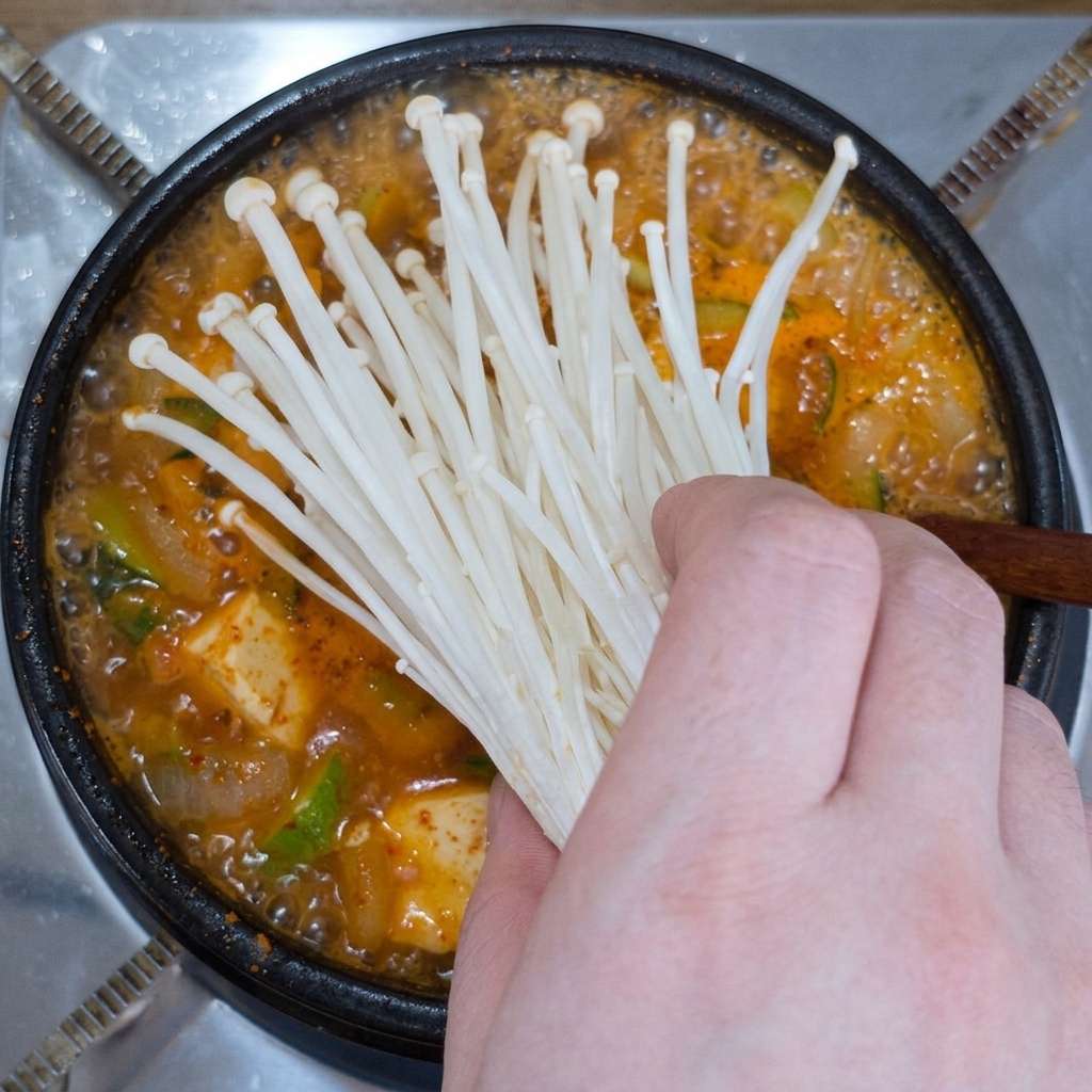 끓고 있는 된장찌개 냄비에 신선한 팽이버섯 한 줌을 얹는 모습.
