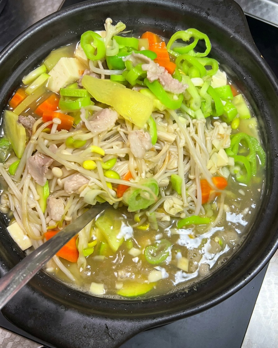 검은색 뚝배기에서 끓고 있는 콩나물, 돼지고기, 푸른 채소 찌개를 숟가락으로 부드럽게 젓는 모습.