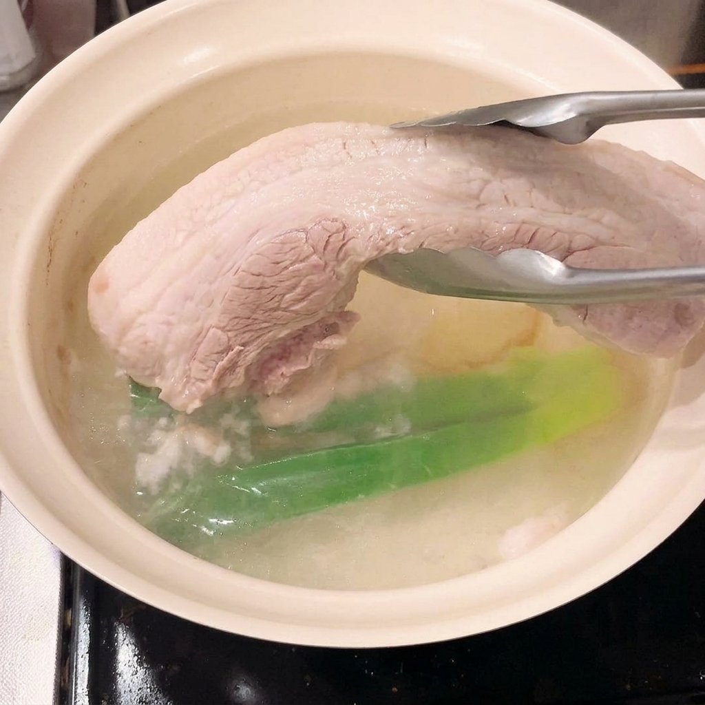 집게로 끓는 물에서 데쳐져 단단해진 삼겹살 덩어리를 들어 올리는 모습.