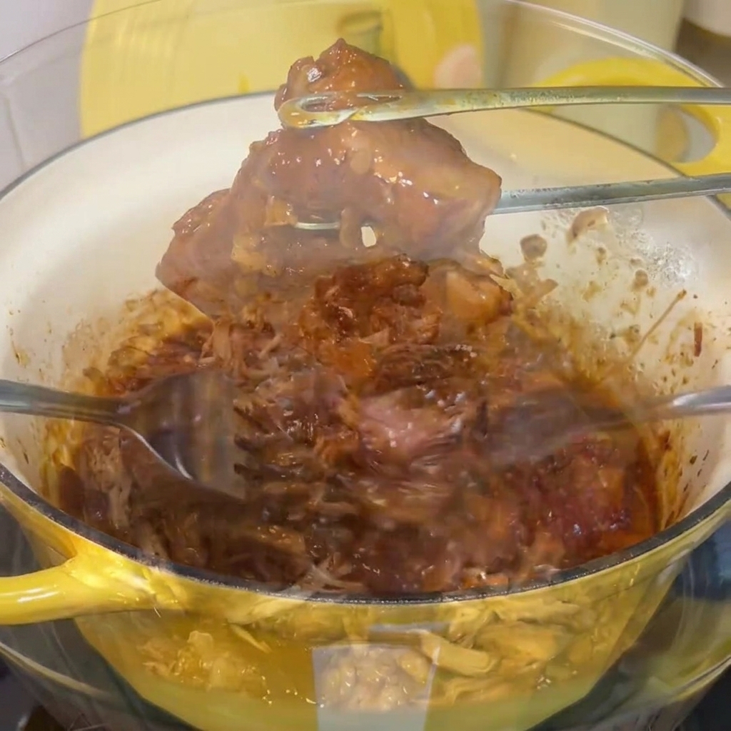 투명 유리 볼에서 포크 두 개를 사용하여 조리된 돼지 어깨살을 잘게 찢는 모습.