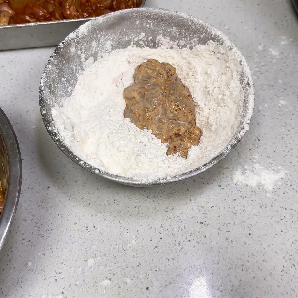 젖은 치킨 조각을 밀가루가 담긴 은색 볼에 넣고 버무리는 모습.