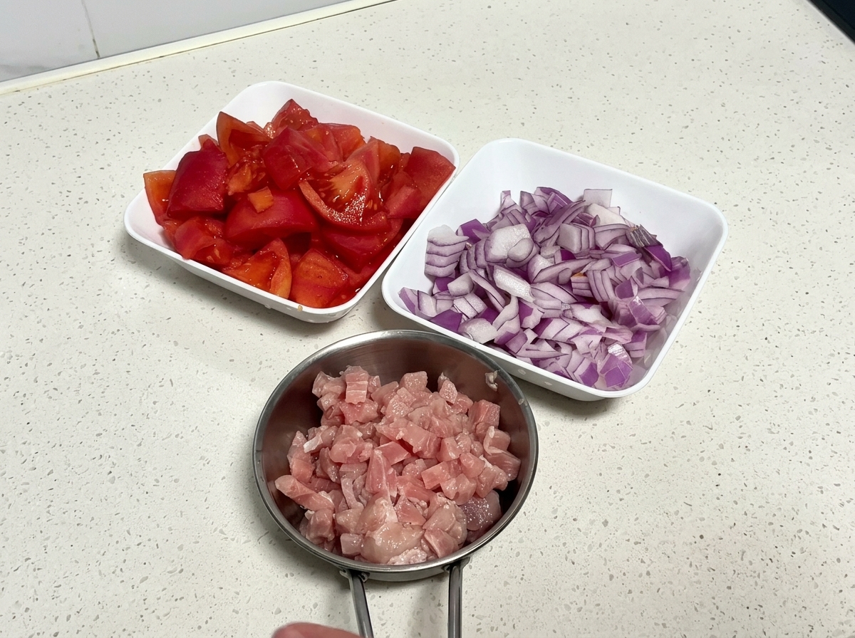 도마 위에 준비된 잘게 썬 토마토, 다진 적양파, 깍둑썰기한 돼지고기가 담긴 세 개의 흰 그릇.