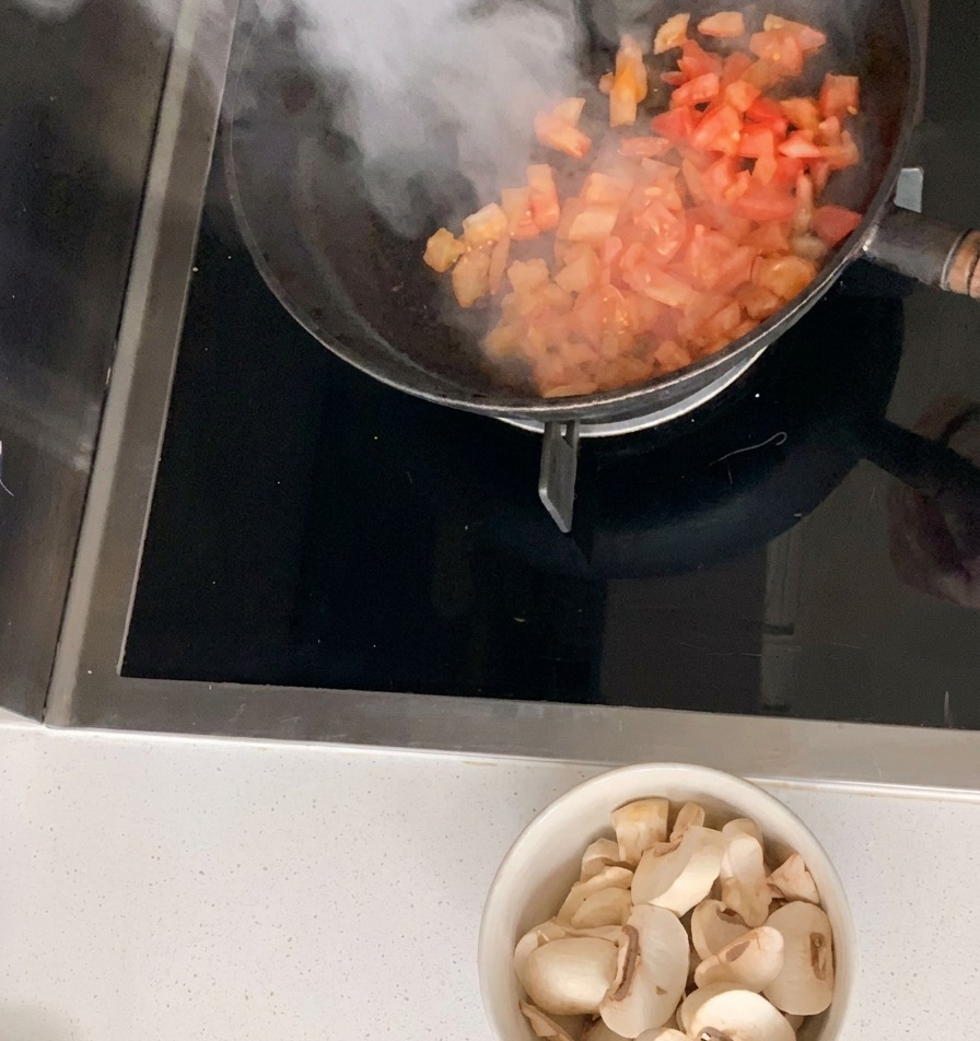 검은색 프라이팬에서 깍둑썰기한 토마토를 볶는 모습. 조리대 아래에는 얇게 썬 양송이버섯이 담긴 그릇이 놓여 있습니다.