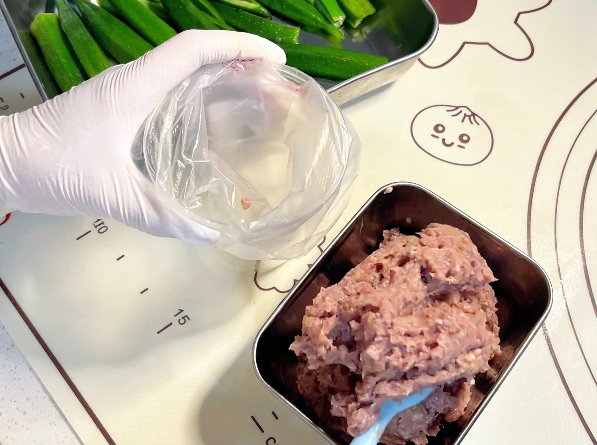 장갑을 낀 손으로 파란색 주걱을 사용해 곱게 갈린 생고기 페이스트를 투명한 플라스틱 짤주머니에 담는 모습.