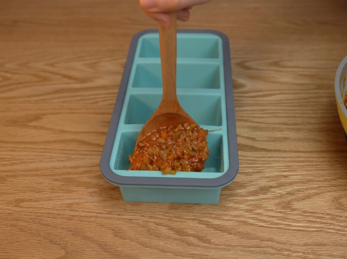 파란색 실리콘 트레이의 각 칸에 걸쭉한 토마토 미트 소스를 숟가락으로 떠 담는 모습.