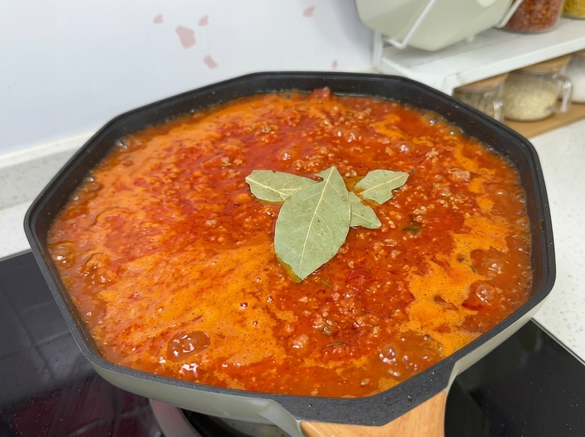 웍 안에서 끓고 있는 토마토 미트 소스에 소금, 설탕, 월계수 잎, 흑후추를 넣고 간을 맞추는 모습.