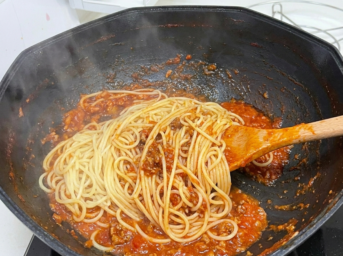 김이 모락모락 나는 검은 웍에서 나무 숟가락을 사용해 삶은 스파게티 면과 진한 토마토 미트 소스를 섞고 있는 모습.