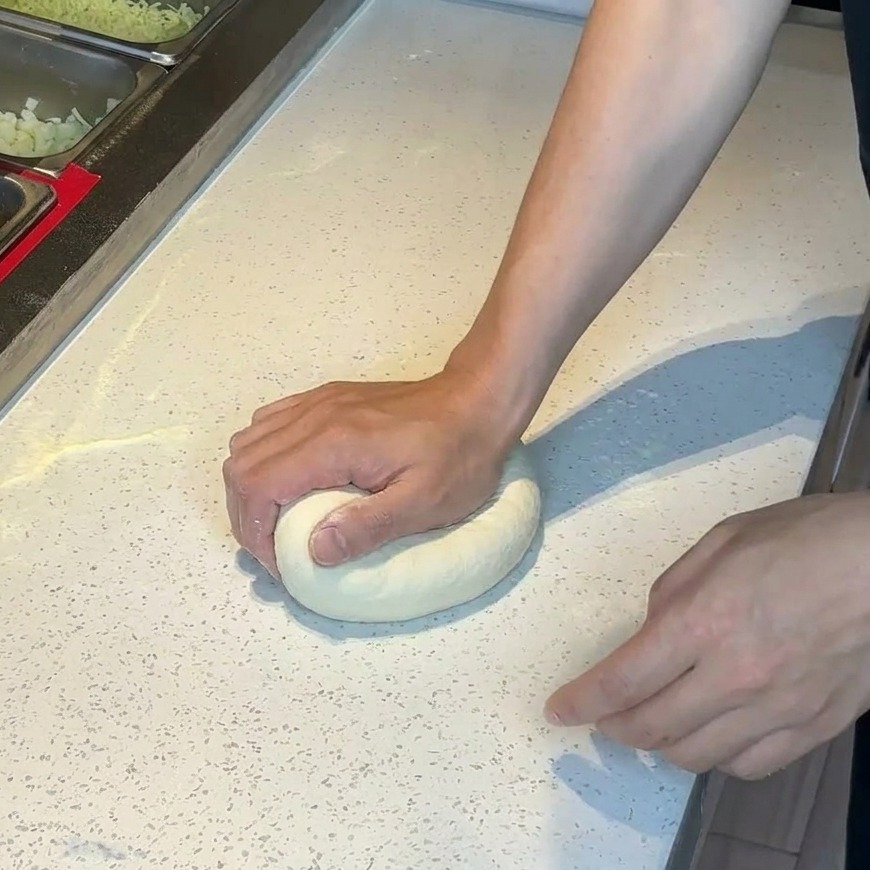 밀가루를 뿌린 조리대에 놓인 피자 도우 반죽을 손으로 누르는 모습.