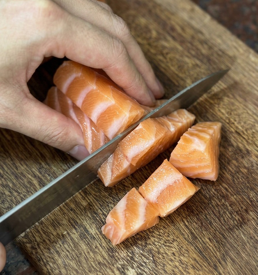 나무 도마 위에서 잘 드는 칼로 신선한 주홍빛 연어를 한입 크기의 정육면체로 썬 모습.
