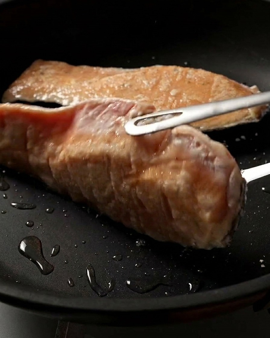 팬 안에서 뒤집히는 연어 필레의 클로즈업 샷으로, 노릇노릇하고 바삭하게 잘 구워진 표면이 보이는 모습.