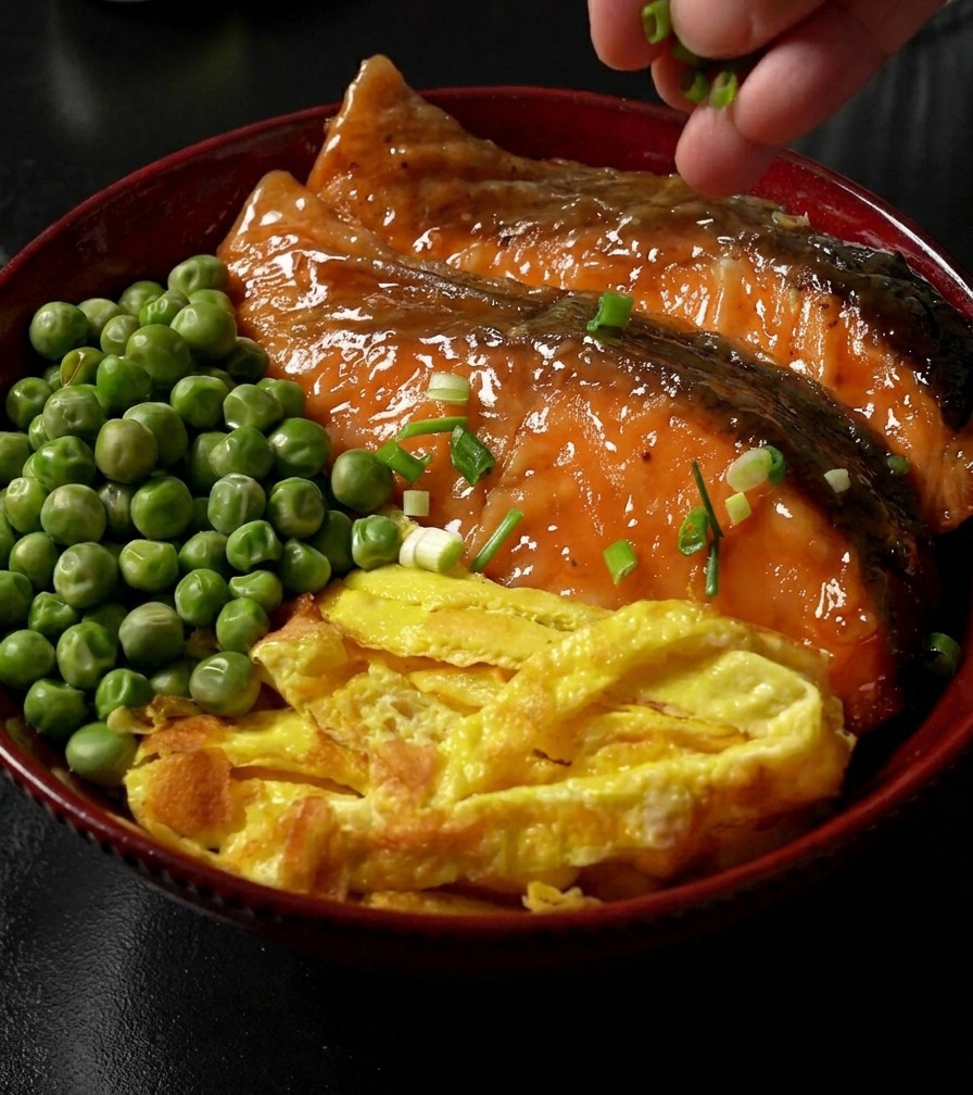 데리야끼 연어 구이, 달걀지단, 완두콩이 올려진 완성된 덮밥 위에 손으로 송송 썬 파를 뿌리는 모습.