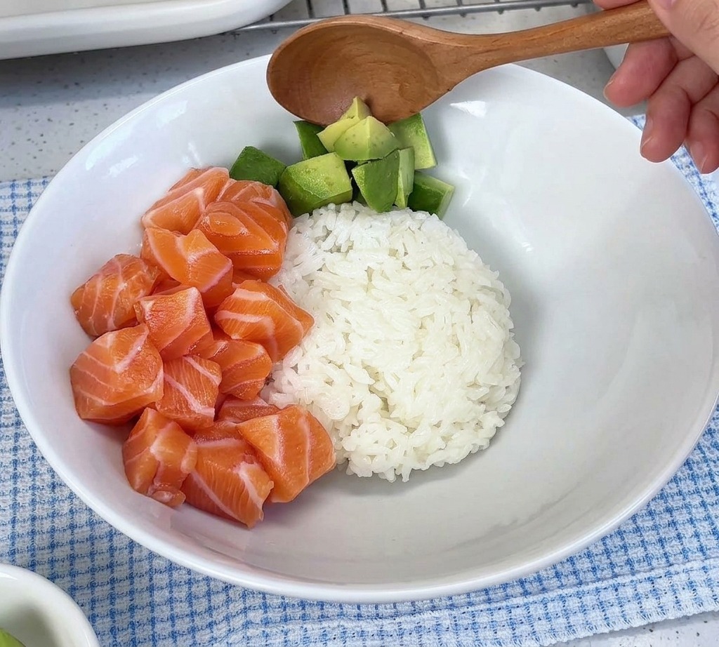 밥과 생연어 조각들이 담긴 하얀 그릇에 깍둑썰기한 아보카도를 숟가락으로 가지런히 얹는 모습.
