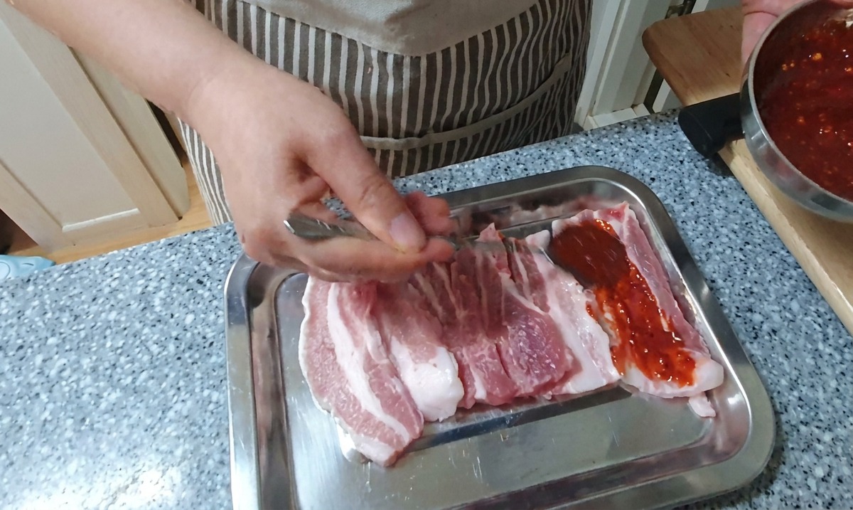 은색 쟁반 위에서 손과 스푼을 이용해 생삼겹살에 진한 빨간색 고추장 양념을 골고루 바르는 모습