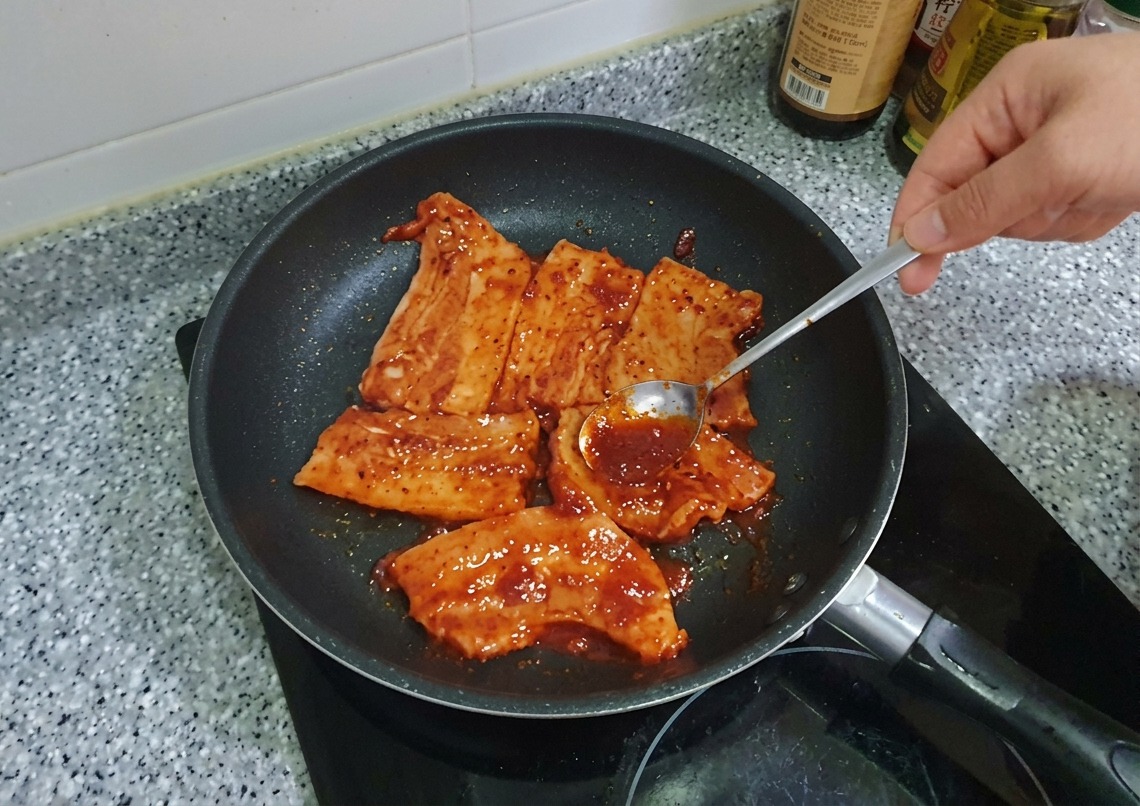 팬 안에서 익어가는 삼겹살 위에 스푼으로 진한 고추장 양념을 추가로 얹어 펴 바르는 모습