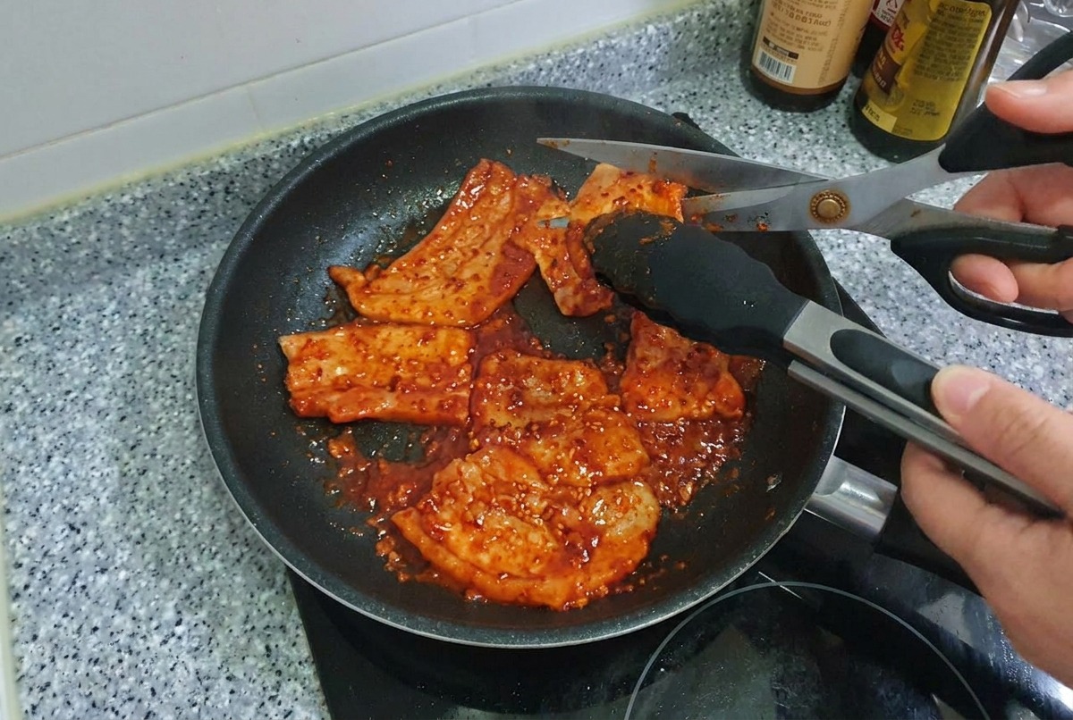 프라이팬 안에서 익어가고 있는 매콤하게 구워진 삼겹살을 요리용 가위로 자르는 모습