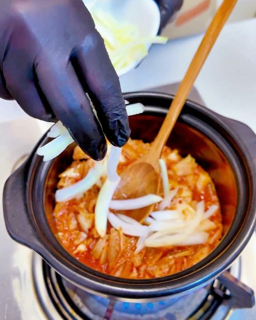 검은 장갑을 낀 손으로 냄비 속 볶은 김치에 채 썬 양파를 넣는 모습