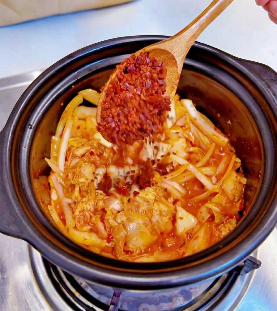 볶은 김치와 양파가 담긴 검은색 냄비에 나무 숟가락으로 굵은 고춧가루를 넣는 모습