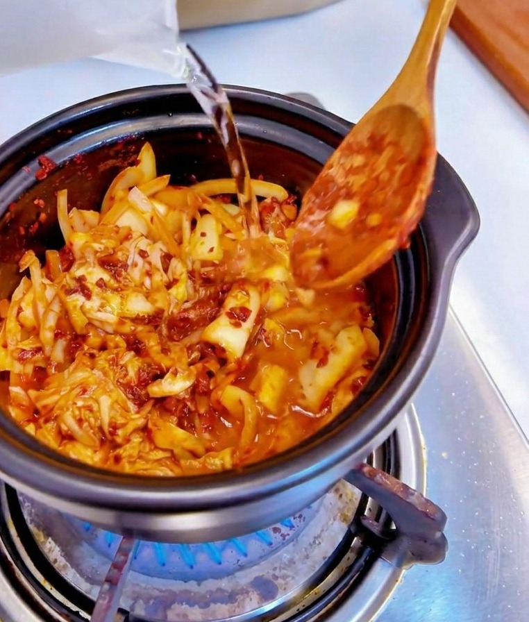 매콤하게 볶아진 김치와 양파가 담긴 검은 냄비에 투명한 계량컵으로 물을 붓는 모습