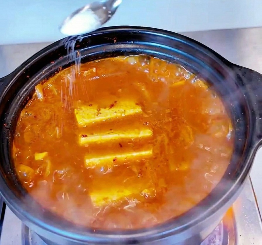 보글보글 끓는 참치김치찌개 위에 작은 금속 숟가락으로 하얀 소금을 뿌려 간을 맞추는 모습