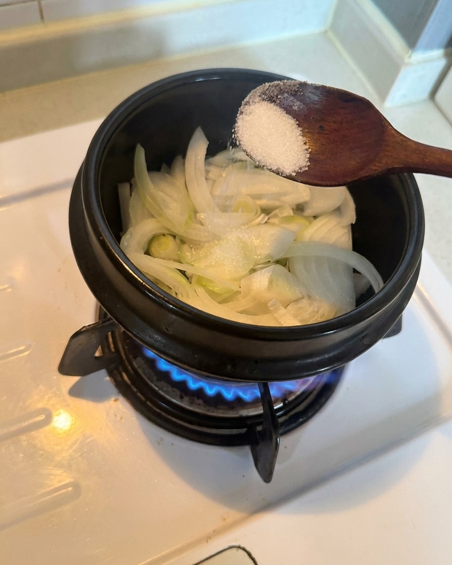 냄비 속 채 썬 양파 위에 흰 설탕 한 스푼을 넣고 불 위에서 볶는 모습