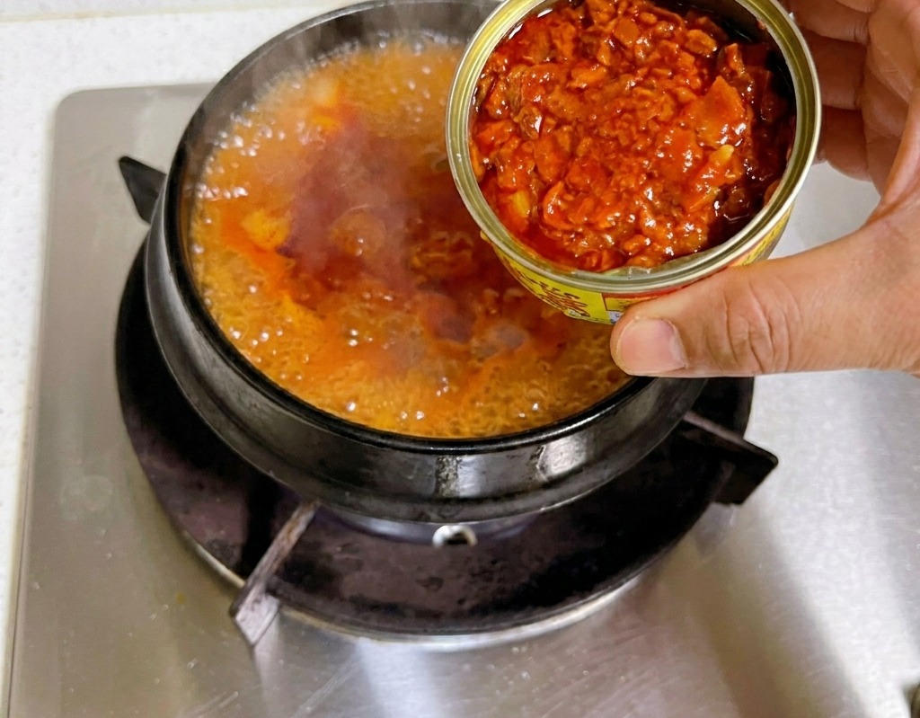 보글보글 끓어오르는 찌개에 참치캔을 기울여 참치 살코기와 기름을 함께 넣는 모습
