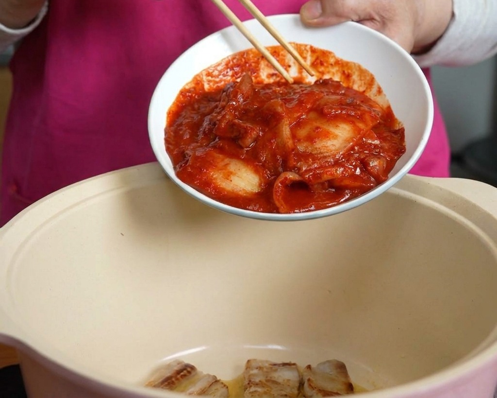 손으로 젓가락을 사용해 매콤한 붉은 양념의 신김치를 삼겹살이 담긴 냄비 안으로 밀어 넣는 모습