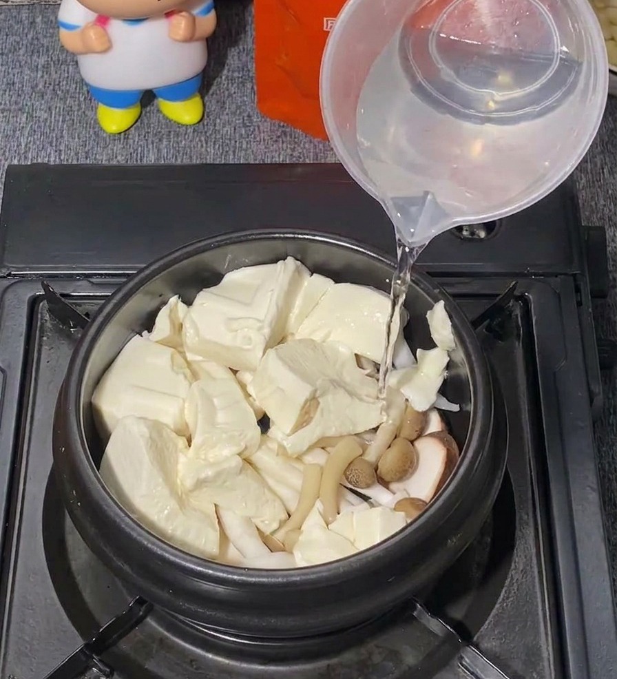 플라스틱 계량컵을 이용해 순두부와 버섯이 담긴 검은색 냄비에 맑은 물을 붓고 있는 모습.