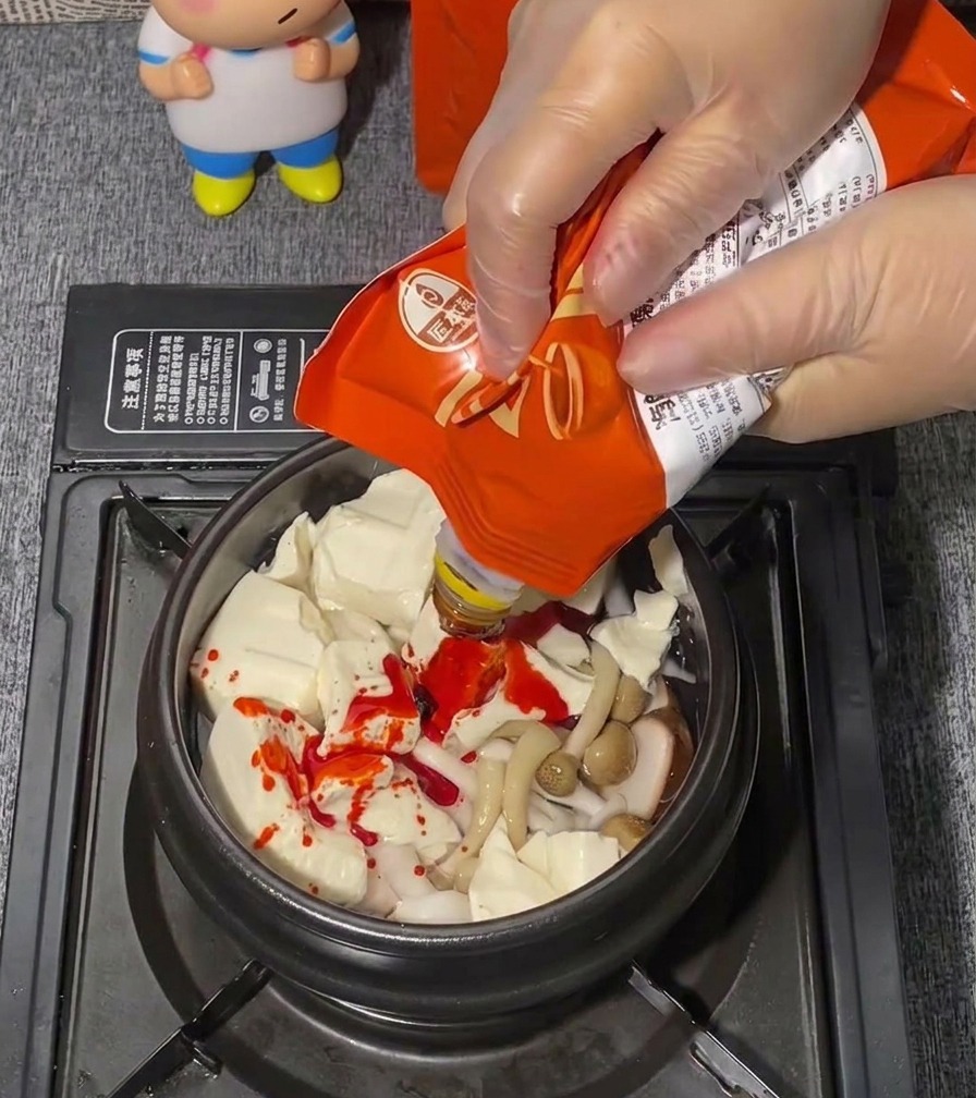 투명한 위생 장갑을 낀 손이 순두부 양념장 파우치를 짜서 순두부 위에 붉은 양념을 얹고 있는 모습.