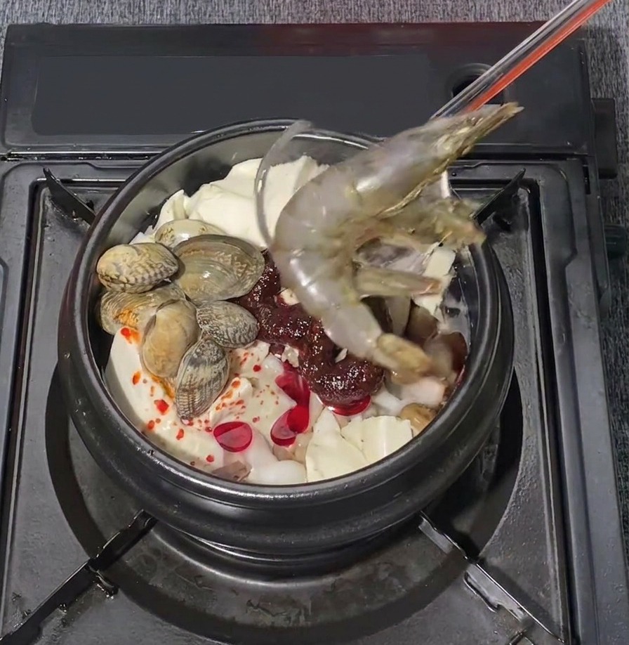 한국식 해물 순두부찌개가 끓고 있는 냄비에 크고 신선한 생새우 두 마리를 조심스럽게 얹는 모습.