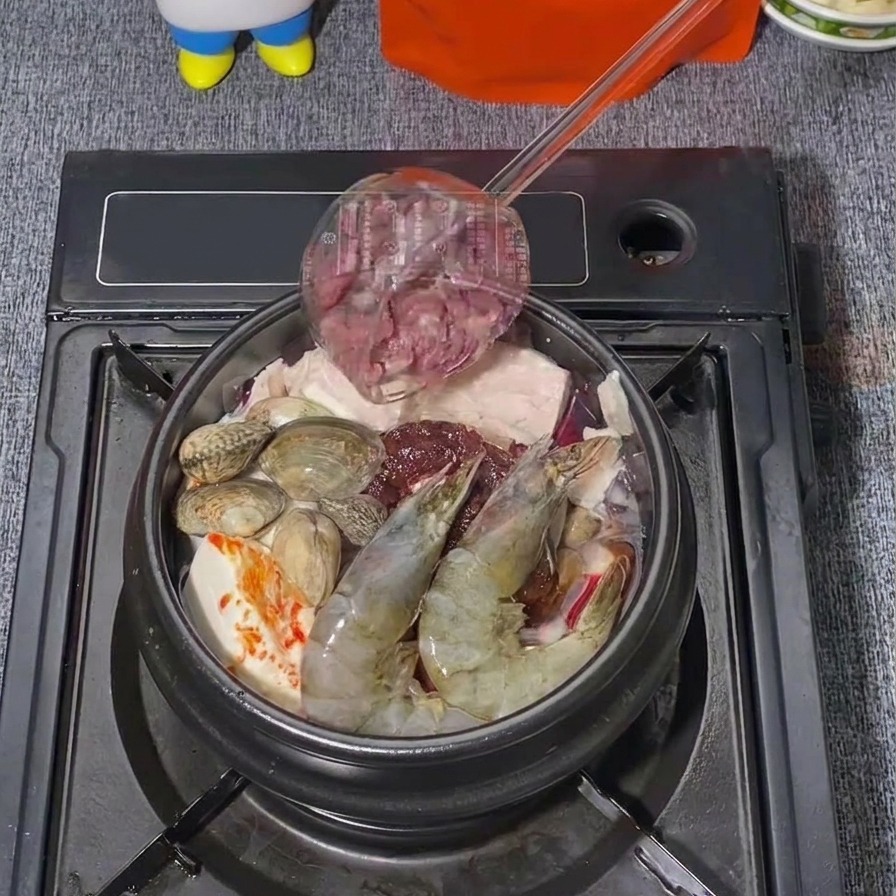 새우와 바지락이 들어간 순두부찌개 냄비 위에 얇게 썬 생소고기를 국자로 얹어 넣는 모습.
