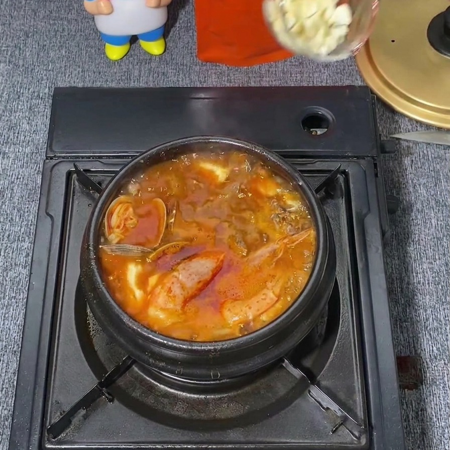 팔팔 끓는 붉은색 매콤한 순두부찌개 중앙에 투명한 국자를 사용해 다진 마늘 한 숟갈을 넣는 모습.