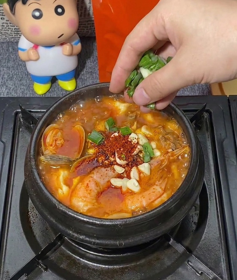 새우, 바지락, 다진 마늘로 가득 차 보글보글 끓고 있는 검은색 뚝배기에 송송 썬 대파를 한 줌 뿌리는 모습.