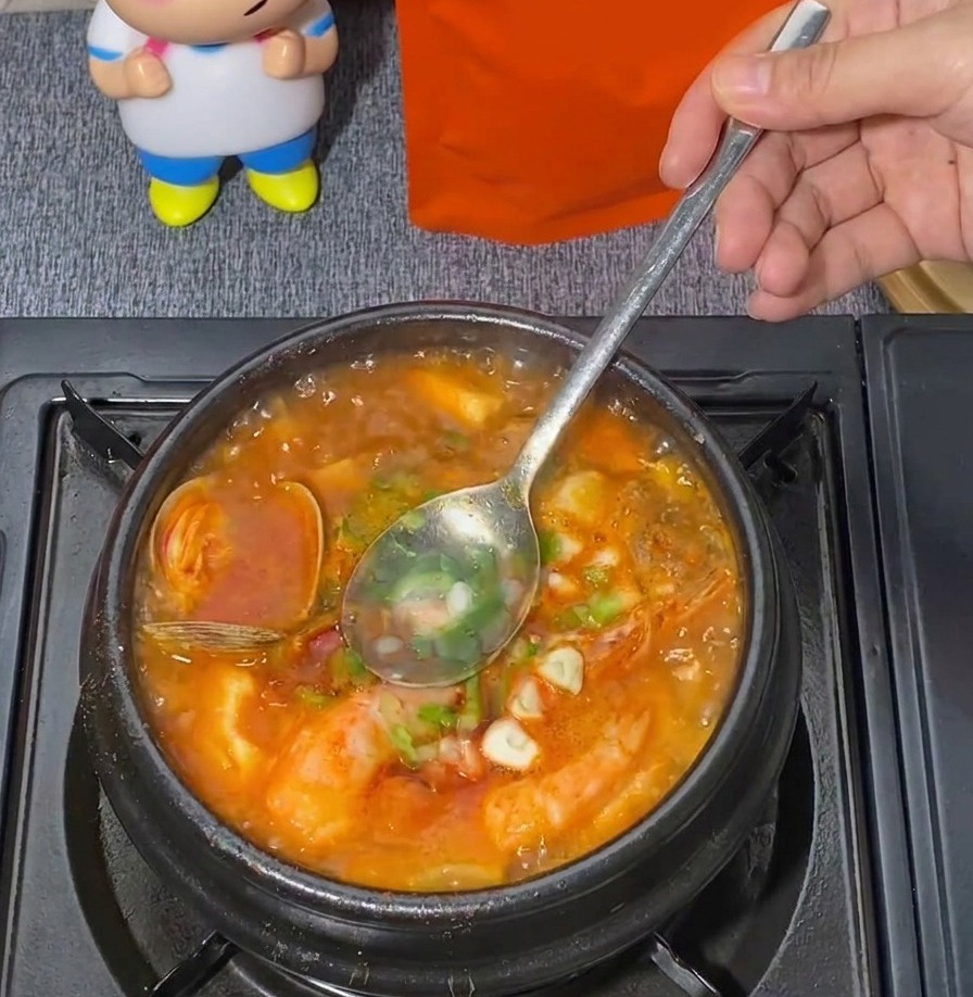 붉은색 해물 순두부찌개가 끓고 있는 냄비에 금속 숟가락을 넣어 송송 썬 대파를 살살 섞어주는 모습.