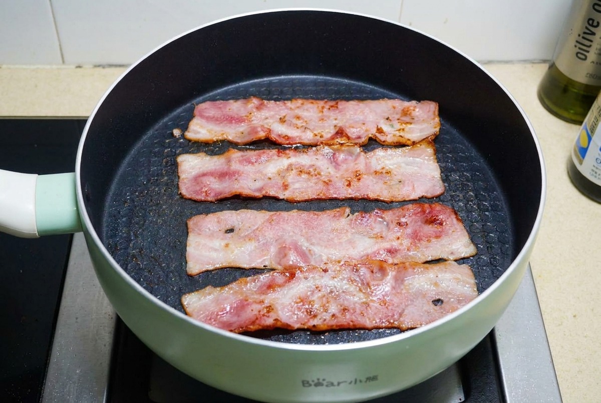 검은색 코팅 팬에서 기름이 보글보글 끓으며 베이컨 네 줄이 노릇하고 바삭하게 구워지고 있는 모습.