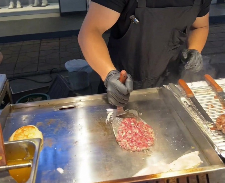 업소용 철판 위에서 금속 뒤집개로 소고기 패티를 뒤집어 구운 적양파 더미 위에 얹는 모습.