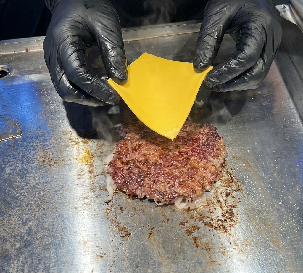 검은색 장갑을 낀 손으로 지글지글 익어가는 노릇한 소고기 패티 위에 노란 체다 치즈 한 장을 조심스럽게 올리는 클로즈업 모습.