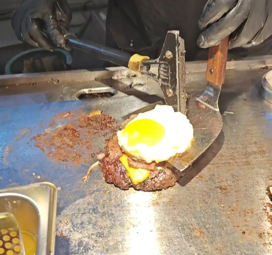 넓은 금속 뒤집개로 소고기 패티, 녹은 치즈, 베이컨, 반숙 달걀 프라이가 층층이 쌓인 버거 스택을 들어올리는 모습.