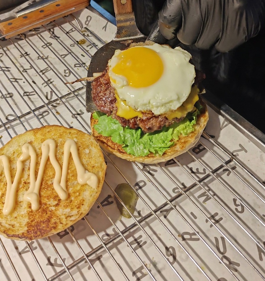 금속 뒤집개를 사용해 치즈가 녹아내린 소고기 패티와 반숙 달걀 프라이 스택을 양상추와 토마토 층 위로 조심스럽게 옮기는 모습.