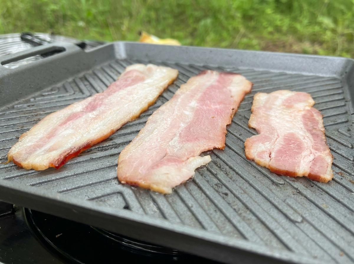 회색 그릴팬 위에서 기름을 지글지글 내며 맛있게 익어가고 있는 긴 베이컨 세 줄.