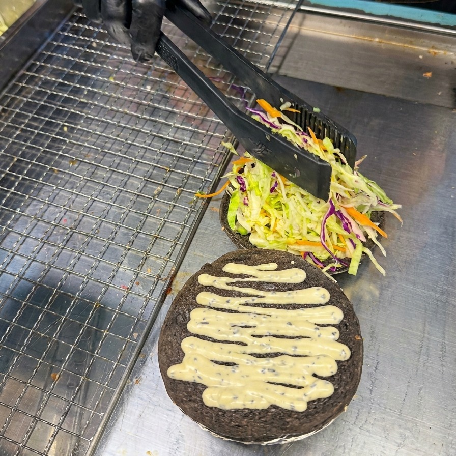 검은색 버거 번 위의 채소 침대 위에 집게로 밝은 붉은색의 도톰한 토마토 슬라이스를 조심스럽게 올리는 모습