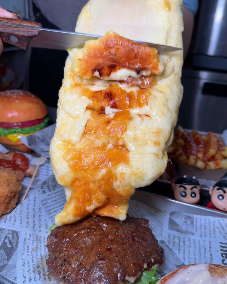칼로 커다란 치즈 휠에서 그을리고 녹은 치즈를 듬뿍 긁어내어 소고기 패티 위에 올리는 모습.