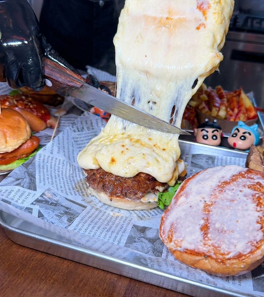 큰 칼로 치즈 휠에서 끓어오르는 두꺼운 치즈를 긁어내어 소고기 버거 패티에 얹는 모습.